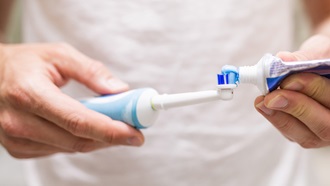 Pessoa com blusa branca coloca pasta de dentes numa escova de dentes elétrica.